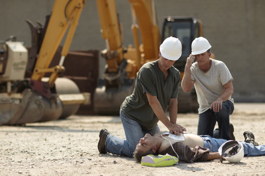 Three men at a construction worksite. One man is on the ground, unconscious and hooked up to an AED. A second man is performing CPR. The third man is making a phone call to emergency personnel.