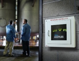 AED in cabinet in foreground with two men talking in an industrial setting in the background